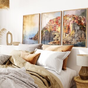 May include: Three framed watercolor paintings of a colorful Italian village on a cliff overlooking the ocean. The paintings are hung on a white wall above a bed with brown bedding and pillows.