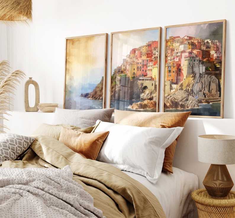 May include: Three framed watercolor paintings of a colorful Italian village on a cliff overlooking the ocean. The paintings are hung on a white wall above a bed with brown bedding and pillows.