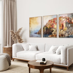 May include: Three framed watercolor paintings of a colorful Italian village on a cliff overlooking the sea. The paintings are hung on a white wall above a white sofa.