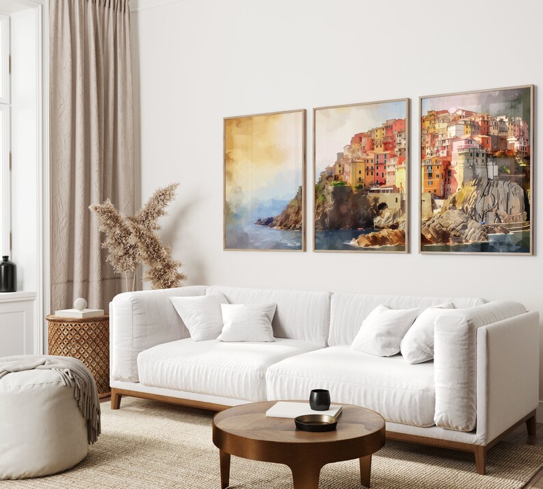 May include: Three framed watercolor paintings of a colorful Italian village on a cliff overlooking the sea. The paintings are hung on a white wall above a white sofa.