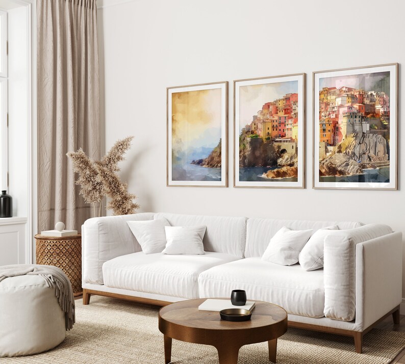 May include: Three framed watercolor paintings of a colorful Italian village on a cliff overlooking the ocean. The paintings are hung on a white wall above a white sofa with pillows.
