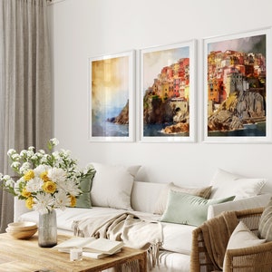 May include: Three framed watercolor paintings of a colorful Italian village on a cliff overlooking the sea. The paintings are in white frames and are hung on a white wall.
