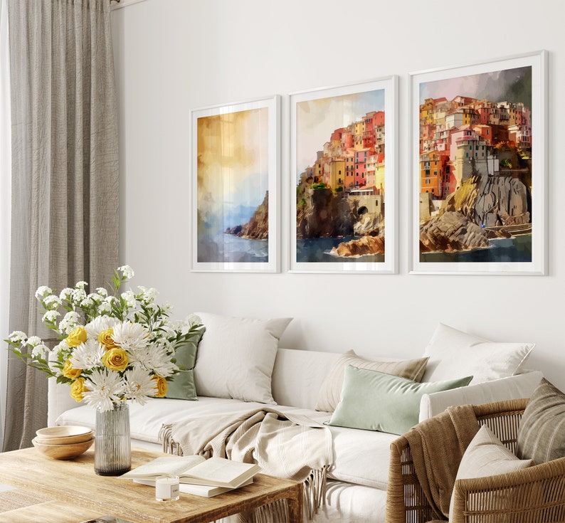 May include: Three framed watercolor paintings of a colorful Italian village on a cliff overlooking the sea. The paintings are in white frames and are hung on a white wall.