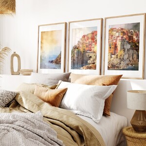May include: Three framed watercolor paintings of a colorful Italian village on a cliff overlooking the ocean. The paintings are hanging on a white wall above a bed with beige linens and pillows.