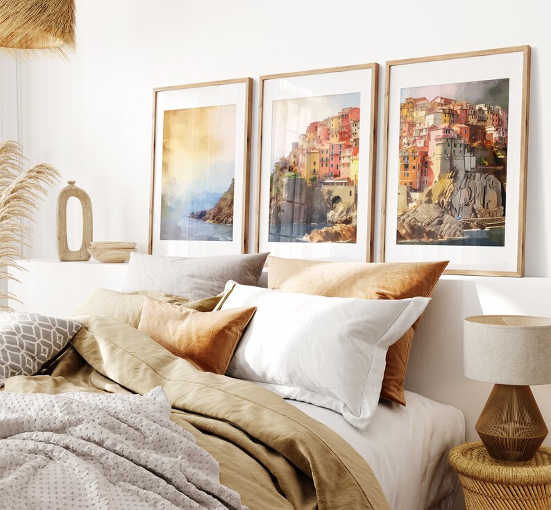 May include: Three framed watercolor paintings of a colorful Italian village on a cliff overlooking the ocean. The paintings are hanging on a white wall above a bed with beige linens and pillows.