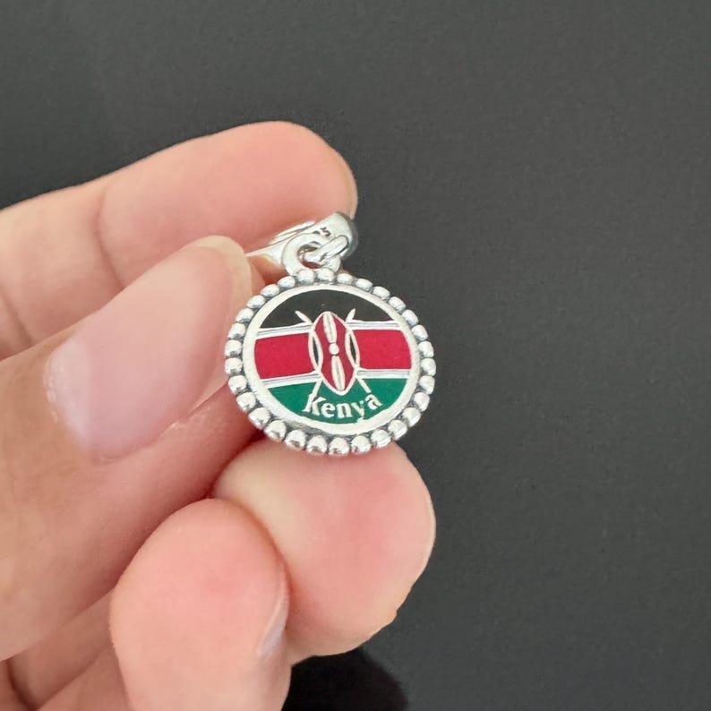 May include: Silver charm with a round enamel design featuring the flag of Kenya. The charm has a beaded border and the word "Kenya" is written below the flag.