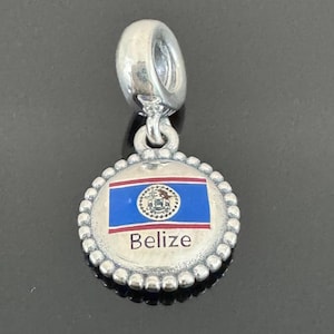 Könnte beinhalten: Silberner Charm mit einem runden Anhänger mit der Flagge von Belize. Das Flaggen-Design umfasst einen blauen Hintergrund, rote Streifen und das Wappen von Belize. Das Wort "Belize" ist in Weiß aufgedruckt. Der Charm hat einen Perlenrand.