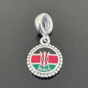 May include: Silver charm with a round enamel pendant featuring the flag of Kenya. The pendant has a black background with a red, white, and green horizontal stripe design. The center of the pendant features a black shield with two spears crossed behind it. The word "Kenya" is written in black letters at the bottom of the pendant.