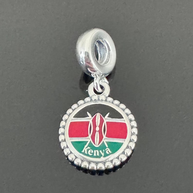 May include: Silver charm with a round enamel pendant featuring the flag of Kenya. The pendant has a black background with a red, white, and green horizontal stripe design. The center of the pendant features a black shield with two spears crossed behind it. The word "Kenya" is written in black letters at the bottom of the pendant.