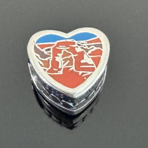 May include: Heart-shaped silver charm with a colorful enamel design. The charm features a landscape scene with red, brown, and white rock formations under a blue sky. The charm is likely a souvenir or jewelry piece.