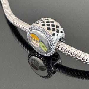 Könnte beinhalten: Ein silbernes Armband mit einem silbernen Anhänger, der einen Stapel von drei bunten Macarons zeigt. Der Anhänger hat ein schwarz-weißes Karomuster und ist mit klaren Zirkonia-Steinen verziert. Der Anhänger ist mit "S925 ALE" gekennzeichnet.