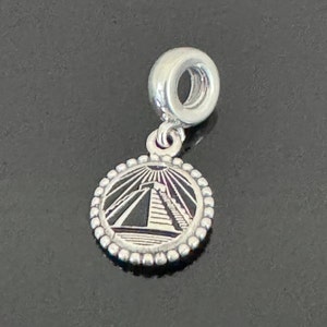 Pode incluir: Berloque de prata com um pingente redondo com uma representação detalhada de uma pirâmide com raios de sol. O berloque tem uma borda com contas e uma argola para prender a uma pulseira ou colar. O berloque é feito de metal prateado brilhante.