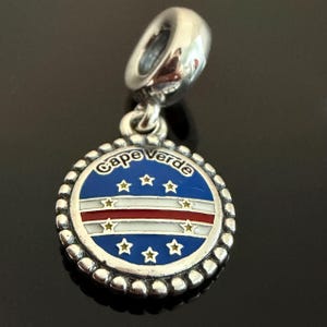 May include: A silver charm featuring a circular pendant with a blue, white, and red enamel design representing the flag of Cape Verde. The text Cape Verde is engraved at the top, surrounded by ten small yellow stars on a blue background.