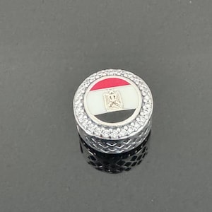 May include: A silver charm with a round, white, black, and red enamel design of the Egyptian flag. The charm is surrounded by a row of clear rhinestones.