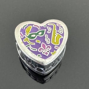 May include: A heart-shaped trinket box with a silver finish. The lid features a purple background with a green mask, yellow saxophone, musical notes, and a fleur-de-lis design. The box is likely a souvenir or decorative item.