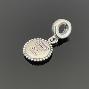 May include: Silver charm with a red and white Texas Tech University logo on a round disc with a beaded edge. The charm has a loop at the top for attaching to a bracelet or necklace.