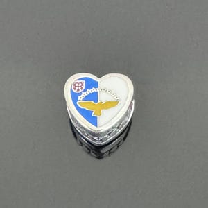 May include: A silver heart-shaped charm with a blue, white, and yellow enamel design. The design features a bird with a red shield on its chest.