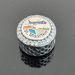 May include: A silver charm with a round, cream-colored center featuring a blue airplane, a map, and the words "St. Maarten." The charm is encircled by clear crystals and has a patterned, openwork design on the sides.