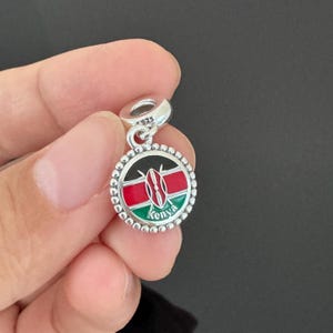 May include: Silver charm with a round, black, red, and green enamel design of the Kenyan flag. The charm has a beaded border and the word "Kenya" is written in white letters below the flag.