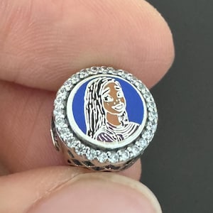 Puede incluir: Un charm de plata con un dise&ntilde;o de esmalte azul y blanco que presenta un personaje de dibujos animados. El charm tiene forma circular con un anillo de piedras transparentes alrededor del borde. El personaje tiene cabello oscuro y una expresi&oacute;n sonriente.
