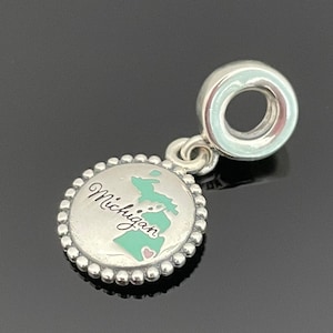Peut inclure: Breloque en argent avec un contour émaillé vert et rose de l'état du Michigan et le mot "Michigan" en cursive. Le charme a une bordure de perles argentées et une boucle argentée pour l'attacher à un bracelet.