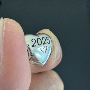 May include: A silver heart-shaped charm with the year "2025" and a heart symbol engraved on it. The word "love" is partially visible on the side. The charm is held between fingers against a dark background.