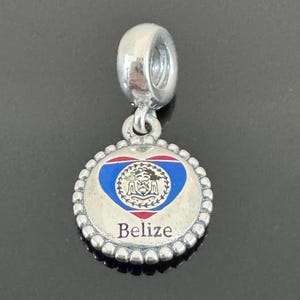 Pode incluir: Um berloque de prata com um pingente redondo com o design da bandeira de Belize. O pingente tem um emblema em forma de coração com o brasão de Belize, rodeado por uma borda de contas, e a palavra "Belize" na parte inferior.