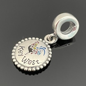 May include: A silver charm with a Key West design. The charm features a colorful rooster illustration and the words "Key West" in a circular design. The charm has a beaded edge and a loop for attaching to a bracelet or necklace.