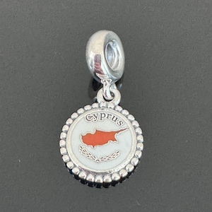 May include: Silver charm with a white and red enamel design of the flag of Cyprus. The charm has a round shape with a beaded edge and the word "Cyprus" written in black lettering.