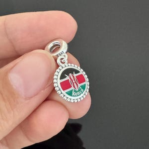 May include: Silver charm with a round enamel pendant featuring the flag of Kenya. The pendant has a black, red, and green design with a shield in the center. The word "Kenya" is written in white letters below the shield.