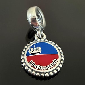 Peut inclure: Un charm en métal argenté avec un pendentif circulaire à bordure perlée. Le centre affiche le drapeau bleu et rouge du Liechtenstein avec une icône de couronne et le texte Liechtenstein imprimé en blanc le long de la courbe inférieure.