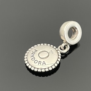 May include: Silver charm with a round, beaded border and the word "Pandora" inscribed on it. The charm has a small loop at the top for attaching to a bracelet or necklace.