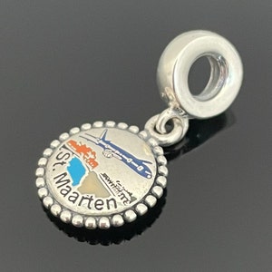 Può includere: Ciondolo in argento con un design in smalto blu, bianco e arancione di un aereo che vola sopra una piccola isola con il testo "St. Maarten".