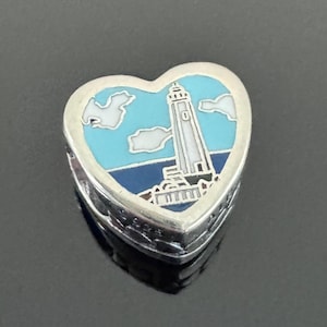 May include: Heart-shaped silver pendant with a lighthouse design. The pendant features a blue sky with white clouds, a white and gray lighthouse, and a blue sea. The edges of the pendant have a textured, brick-like pattern.