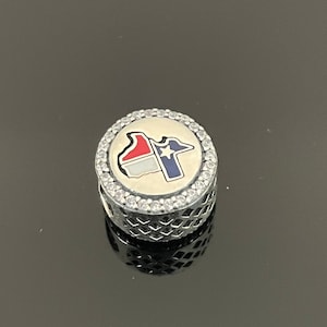 May include: Silver charm with a white, red, and blue enamel outline of the state of Texas with a white star in the blue section. The charm is surrounded by a circle of clear cubic zirconia stones.