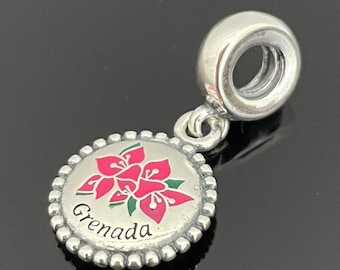 May include: Silver charm with a round, white enamel disc featuring a pink flower design and the word "Grenada". The charm has a silver loop for attaching to a bracelet.
