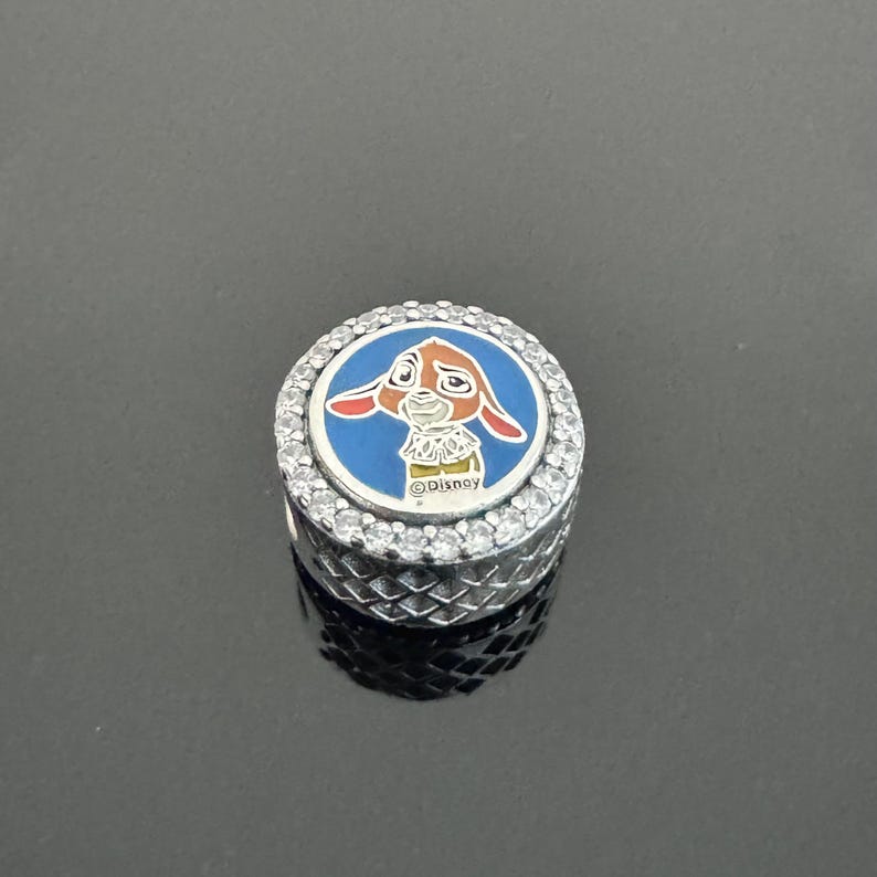 Puede incluir: Un charm de plata con un dise&ntilde;o de esmalte azul y blanco con un conejo de dibujos animados. El charm est&aacute; rodeado de piedras redondas y transparentes y tiene la palabra "Disney" impresa.