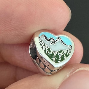 Peut inclure: Un charm en argent en forme de cœur avec un motif de paysage de montagne. Le charm présente un ciel bleu, des montagnes blanches et des arbres verts. Le charm est marqué "S925".