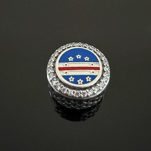 May include: A round silver charm bead featuring a central circular inlay with the flag of Cape Verde, displaying blue, white, and red horizontal stripes with a ring of ten gold-coloured stars, surrounded by a border of small clear crystals.