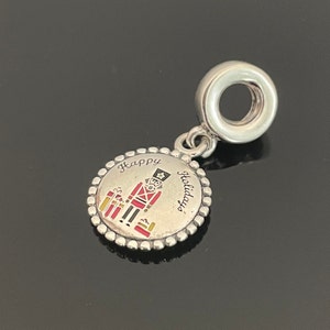 Pode incluir: Berloque de prata com um design de quebra-nozes vermelho e dourado e o texto "Happy Holidays".
