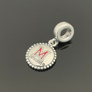 May include: Silver charm with a red "M" on a white background. The charm has a beaded edge and says "UM" on the bottom.