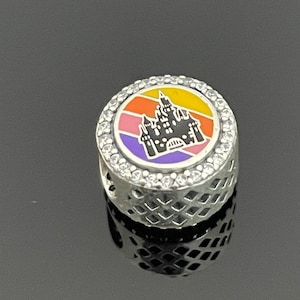 May include: A silver charm with a colorful enamel design of a castle silhouette against a sunset backdrop. The charm is surrounded by a row of clear cubic zirconia stones.