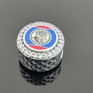 May include: A silver charm with a Belize flag design. The charm has a circular design with the Belize flag in the centre, surrounded by clear crystals. The sides of the charm have a diamond pattern.