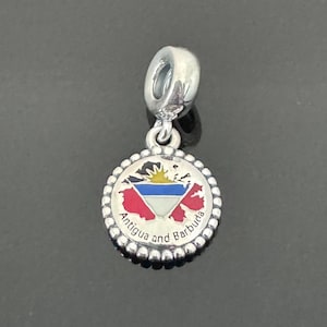 May include: A silver charm with a round pendant featuring the Antigua and Barbuda flag. The flag design includes red, blue, and white elements, with the words "Antigua and Barbuda" around the edge. The charm has a loop for attaching to a bracelet or necklace.