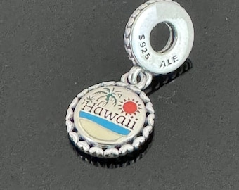 Op de afbeelding: Zilveren bedel met een ronde, witte emaille hanger met een palmboom en het woord "Hawaii" in blauwe letters. De bedel heeft een zilveren kralenrand en een rode zon. De achterkant van de bedel heeft de inscriptie "S925 ALE".