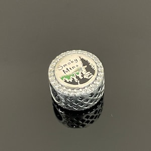 May include: Silver charm with a black and green image of a mountain range with the text "Smoky Mtns" and a clear crystal border.