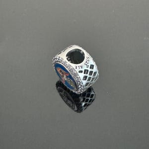 Puede incluir: Un charm de plata con un dise&ntilde;o azul y naranja. El charm tiene forma cuadrada con un patr&oacute;n de diamantes en los lados y peque&ntilde;as piedras transparentes alrededor del dise&ntilde;o azul. La palabra "ALE" es visible en el lateral.