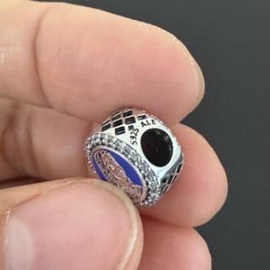 Puede incluir: Un charm de plata con un retrato azul y blanco, rodeado de cristales transparentes. El charm tiene un patr&oacute;n de cuadros negros y plateados y el texto "ALE S925".