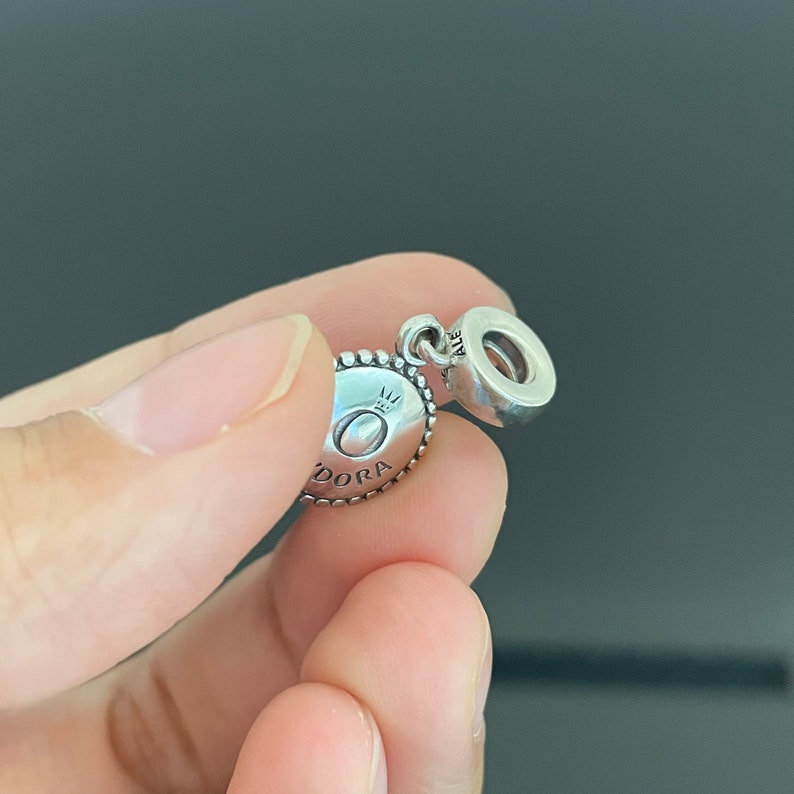 May include: Silver charm with a round, textured design featuring the word "Pandora" and a crown. The charm has a silver loop for attaching to a bracelet.