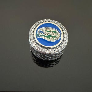 May include: A round silver charm bead featuring a central blue enamel inlay with a green and gold alligator head graphic. The outer edge is encrusted with clear round stones, and the side of the bead has a textured, crisscross metal pattern.